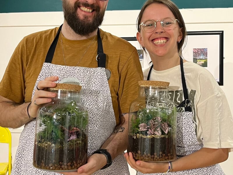 Two smiling people are holding glass terrariums made at Common Room Social's terrarium workshop